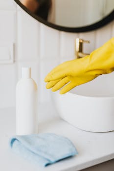 Crop unrecognizable person in yellow protective cleaning glove reaching for detergent bottle and microfiber cloth on washbasin tabletop